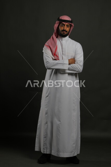 مفهوم الاناقه و الرجولة ،الوقوف باستقامة وثقه بالنفس، بورتريه لشاب وسيم عربي خليجي سعودي مبتسم يرتدي الشماغ و الزي التقليدي، النظر إلى الكاميرا بإيماءات أيدي متقاطعه، الاهتمام بالمظهر الخارجي، صورة الجسم بالطول الكامل، خلفية رمادية