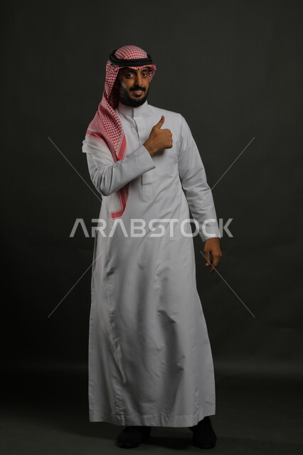 رفع اصبع الابهام للأعلى بإيماءات علامة الجودة, حركات الاعجاب والقبول, بورتريه لرجل عربي خليجي سعودي مبتسم يرتدي الثوب والشماغ التقليدي ينظر إلى الكاميرا بإشارات الموافقه، صورة الجسم بالطول الكامل، خلفية رمادية