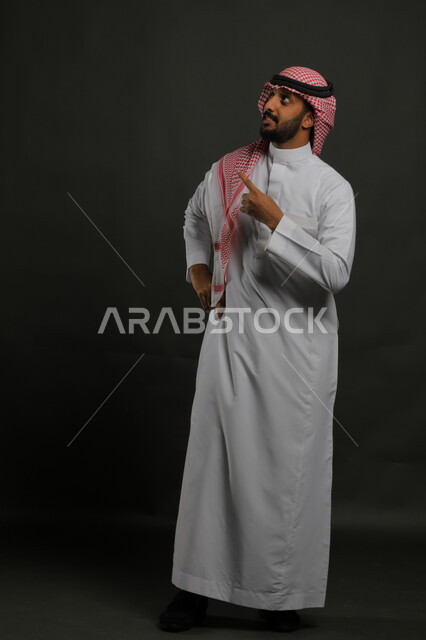 تعابير تدل على الحيرة في اختيار بعض الاشياء، الاشارة إلى مكان معين بإيماءات السرور ,تعابير التأكد والتحديد, بورتريه لرجل عربي خليجي سعودي مبتسم يرتدي الشماغ و الزي التقليدي يرفع إصبعه السبابة للاختيار وينظر إلى شيء ما, صورة للجسم بالكامل, خلفية رمادية