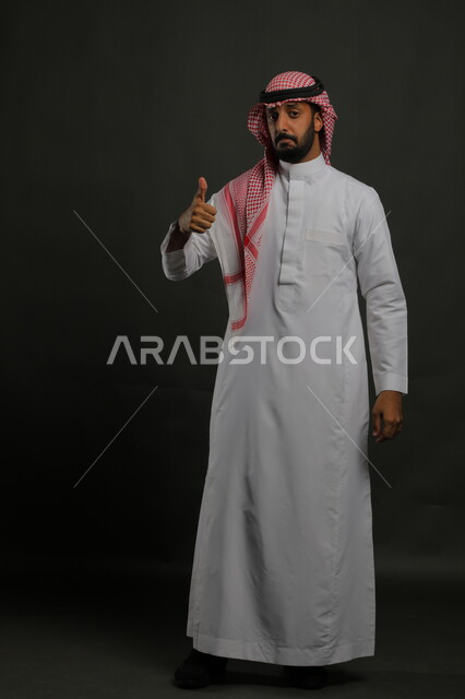 التعبير عن الرأي بالاعجاب والقبول, بورتريه لرجل عربي خليجي سعودي يرتدي الثوب والشماغ التقليدي يرفع اصبع الابهام للأعلى بإيماءات الامتياز و علامة الجودة, النظر إلى الكاميرا بإشارات الموافقه، صورة الجسم بالطول الكامل، خلفية رمادية