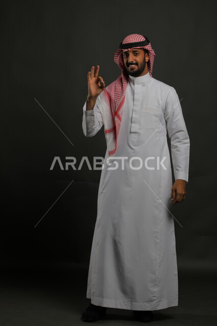 النظر إلى الكاميرا بإشارات الموافقه، حركات الاعجاب والقبول, بورتريه لرجل عربي خليجي سعودي مبتسم يرتدي الثوب والشماغ التقليدي يرفع اصبع الابهام للأعلى بإيماءات علامة الجودة, صورة الجسم بالطول الكامل، خلفية رمادية