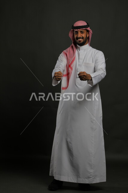 Joy and pleasure in winning business deals, movements indicating victory, profit, and completion of tasks, gestures of victory and enthusiasm, portrait of a smiling Saudi Gulf Arab young man wearing a shemagh and traditional dress, raising his hands forward interactively, full-length body portrait, gray background