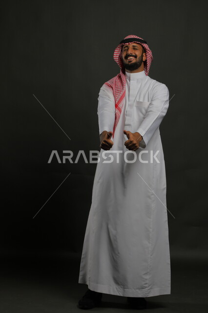 السرور والفرح بتحقيق الاهداف و نيل المراد ,النظر إلى الكاميرا بإشارات الموافقة والاعجاب والقبول، بورتريه لرجل عربي خليجي سعودي مبتسم يرتدي الثوب والشماغ التقليدي يرفع أصابع الابهام بإيماءات علامة الجودة, صورة الجسم بالطول الكامل، خلفية رمادية