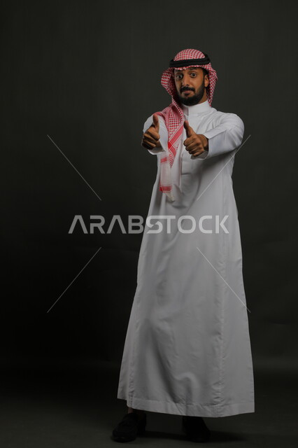 الحماس بالقبول والاعجاب , بورتريه لرجل عربي خليجي سعودي يرتدي الثوب والشماغ التقليدي يرفع أصابع الابهام للأعلى بإيماءات علامة الجودة, النظر إلى الكاميرا بإشارات الموافقه، صورة الجسم بالطول الكامل، خلفية رمادية