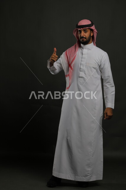 حركات الاعجاب والقبول, بورتريه لرجل عربي خليجي سعودي يرتدي الثوب والشماغ التقليدي يرفع اصبع الابهام للأعلى بإيماءات علامة الجودة, النظر إلى الكاميرا بإشارات الموافقه، صورة الجسم بالطول الكامل، خلفية رمادية