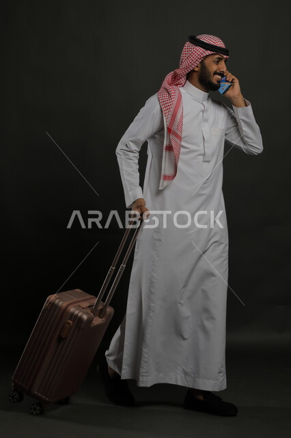 التواصل مع الأهل والأصدقاء, إجراء مكالمات هاتفية عبر الجوال, بورتريه لشاب عربي سعودي خليجي مبتسم يرتدي الثوب التقليدي والشماغ يمسك بيده الهاتف المحمول ويقوم بجر حقيبة السفر, حجز مباشر عبر تطبيقات وبرامج السياحة, صورة الجسم بالطول الكامل, خلفية رمادية