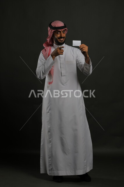 إيماءات الاقناع والإعجاب بطرق الشراء الحديثة, استخدام بطاقات الائتمان البنكية, بورتريه لرجل عربي خليجي سعودي يرتدي الثوب والشماغ التقليدي يمسك بطاقة بيضاء فارغة, الوقوف باستقامة والنظر إلى الكاميرا بابتسامة وسرور, صورة الجسم بالكامل, خلفية رمادية