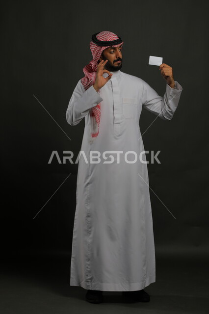 عرض بطاقة عمل تعريفية شخصية بإيماءات الموافقة والجوده و الإعجاب, بورتريه لرجل عربي خليجي سعودي يرتدي الثوب التقليدي والشماغ يمسك بطاقة بيضاء فارغة, الشراء والدفع بطريقة بطاقات الائتمان البنكية, صورة الجسم بالطول الكامل, خلفية رمادي