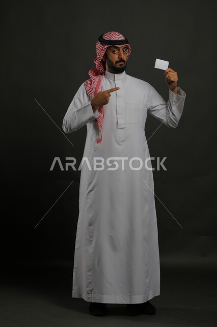 إيماءات الاقناع والإعجاب بطرق الشراء الحديثة, استخدام بطاقات الائتمان البنكية, بورتريه لرجل عربي خليجي سعودي يرتدي الثوب والشماغ التقليدي يمسك بطاقة بيضاء فارغة, الوقوف باستقامة والنظر إلى الكاميرا, صورة الجسم بالكامل, خلفية رمادية