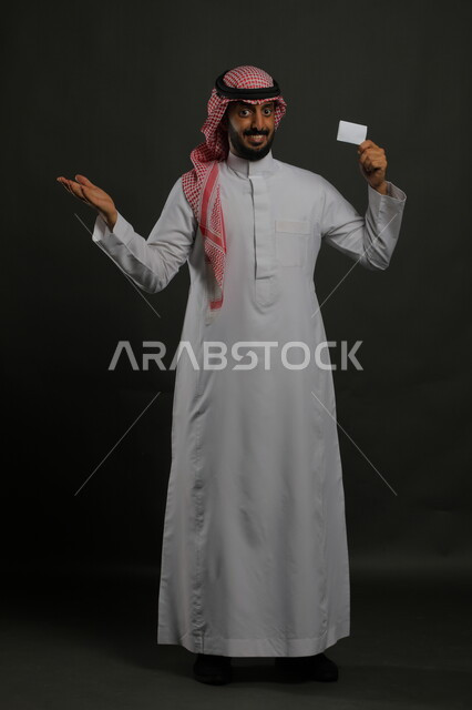 الوقوف باستقامة والنظر إلى الكاميرا وعرض بطاقة تعريفية شخصية, الإعجاب والسرور بطرق الشراء الحديثة, استخدام بطاقات الائتمان البنكية, بورتريه لرجل عربي خليجي سعودي يرتدي الثوب والشماغ التقليدي يمسك بطاقة بيضاء فارغة, صورة الجسم بالكامل, خلفية رمادية