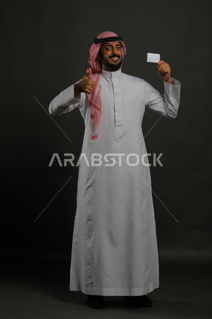 عرض بطاقة عمل تعريفية شخصية بإيماءات الموافقة والجوده و الإعجاب, بورتريه لرجل عربي خليجي سعودي مبتسم يرتدي الثوب التقليدي والشماغ يمسك بطاقة بيضاء فارغة, الشراء والدفع بطريقة بطاقات الائتمان البنكية, صورة الجسم بالطول الكامل, خلفية رمادي