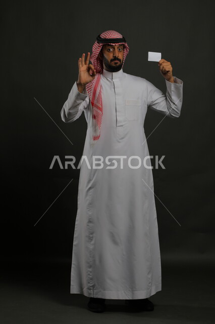 عرض بطاقة عمل تعريفية شخصية بإيماءات الموافقة والجوده و الإعجاب, بورتريه لرجل عربي خليجي سعودي يرتدي الثوب التقليدي والشماغ يمسك بطاقة بيضاء فارغة, الشراء والدفع بطريقة بطاقات الائتمان البنكية, صورة الجسم بالطول الكامل, خلفية رمادي