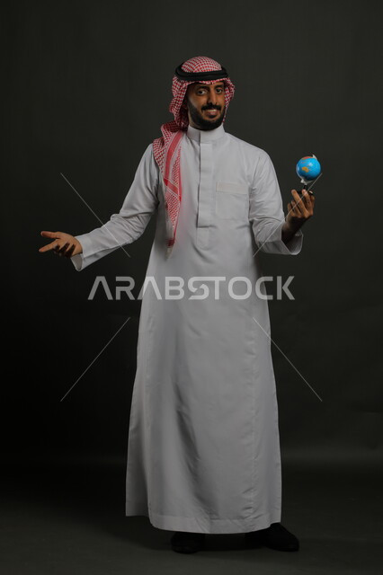 الاستفسار والسؤال عن وجهة سياحية معينة, مفهوم البحث والاستكشاف حول العالم, النظر إلى الكاميرا بإيماءات الحيرة, بورتريه لرجل عربي خليجي سعودي مبتسم يرتدي الثوب التقليدي والشماغ يحمل بيده مجسم الكرة الارضية, صورة الجسم بالطول الكامل, خلفية رمادية