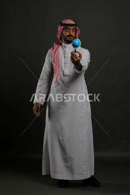 عرض مجسم الكرة الأرضية, بورتريه لرجل عربي خليجي سعودي يرتدي الثوب التقليدي والشماغ يقف باستقامة, مفهوم البحث واستكشاف القارات, القيام برحلة سياحية حول العالم, صورة الجسم بالطول الكامل, خلفية رمادية