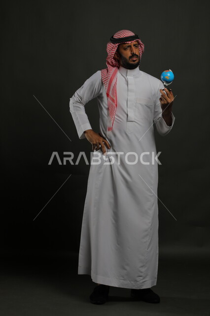 شرود الذهن والتفكير في أمر معين, القيام برحلات سياحية استكشافية حول العالم, بورتريه لرجل عربي خليجي سعودي يمسك مجسم الكرة الأرضية بإيماءات الحيرة بتحديد مكان ما, صورة الجسم بالطول الكامل, خلفية رمادية