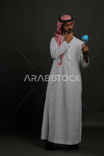 الحيرة والتفكير في أمر ما, القيام برحلات سياحية استكشافية حول العالم, بورتريه لرجل عربي خليجي سعودي يمسك مجسم الكرة الأرضية بإيماءات تحديد مكان معين على الخريطة, صورة الجسم بالطول الكامل, خلفية رمادية