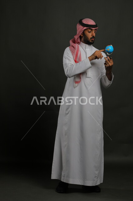 إيماءات إيجاد مكان معين على الخريطة والنظر إلى أحد القارات, مفهوم البحث والاستكشاف حول العالم, بورتريه لرجل عربي خليجي سعودي يرتدي الثوب التقليدي والشماغ يحمل بيده مجسم الكرة الارضية, صورة الجسم بالطول الكامل, خلفية رمادية