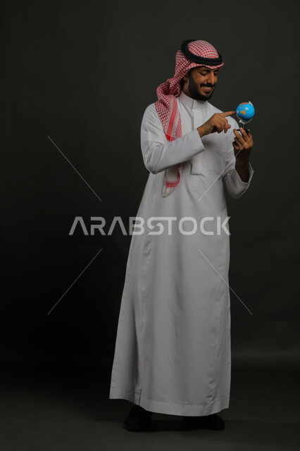 مفهوم البحث والاستكشاف حول العالم, الاشارة إلى مكان معين على الخريطة والنظر إلى أحد القارات, بورتريه لرجل عربي خليجي سعودي مبتسم يرتدي الثوب التقليدي والشماغ يحمل بيده مجسم الكرة الارضية, صورة الجسم بالطول الكامل, خلفية رمادية