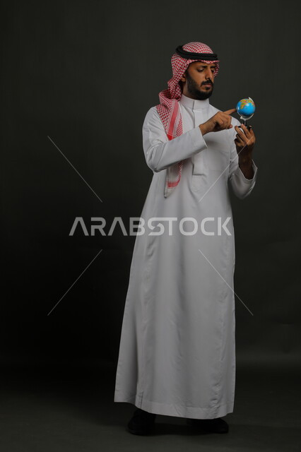 ايماءات التركيز ومفهوم البحث والاستكشاف حول العالم, الاشارة إلى مكان معين على الخريطة والنظر إلى أحد القارات, بورتريه لرجل عربي خليجي سعودي يرتدي الثوب التقليدي والشماغ يحمل بيده مجسم الكرة الارضية, صورة الجسم بالطول الكامل, خلفية رمادية