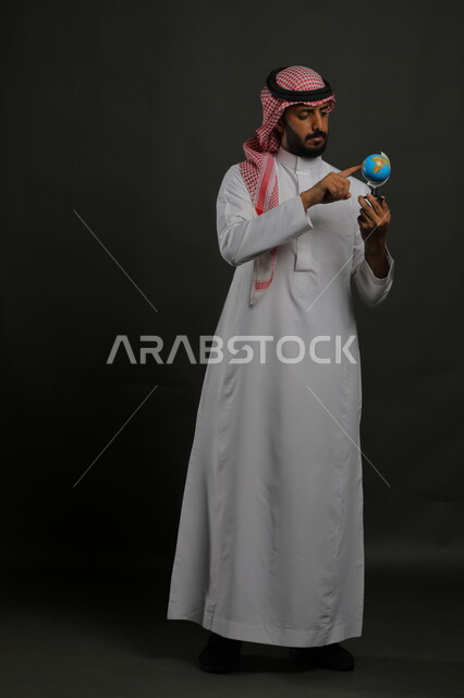 The concept of searching and exploring around the world, pointing to a specific place on the map and looking at one of the continents, a portrait of a Saudi Gulf Arab man wearing the traditional thobe and shemagh, holding a globe in his hand, full-length body image, gray background