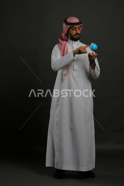 مفهوم البحث والاستكشاف حول العالم, الاشارة إلى مكان معين على الخريطة والنظر إلى أحد القارات, بورتريه لرجل عربي خليجي سعودي يرتدي الثوب التقليدي والشماغ يحمل بيده مجسم الكرة الارضية, صورة الجسم بالطول الكامل, خلفية رمادية