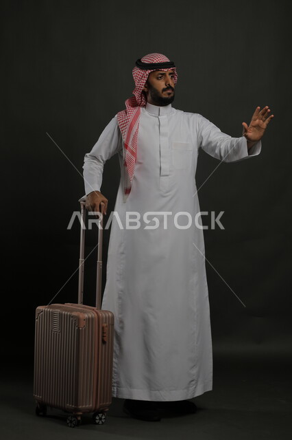 رفع اليد بطلب عدم التدخل والمساعدة, بورتريه لرجل عربي خليجي سعودي يرتدي الثوب التقليدي والشماغ يمسك حقيبة السفر بإيماءات الانزعاج, الصمت والتوقف عن الكلام وعدم القيام بفعل معين, صورة الجسم بالكامل, خلفية رمادية