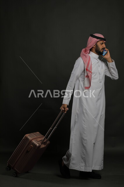 الاستفسار والسؤال عن أمر ما بإيماءات الغضب والقلق, الاستفهام عن خدمات العملاء للرحلة السياحية, بورتريه لرجل عربي خليجي سعودي يرتدي الثوب التقليدي والشماغ يقوم بإجراء مكالمة هاتفية عبر الهاتف المحمول, صورة الجسم بالطول الكامل, خلفية رمادية