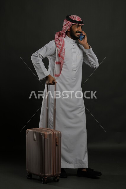 التواصل مع الأهل والأصدقاء, إجراء مكالمات هاتفية عبر الجوال, بورتريه لشاب عربي سعودي خليجي مبتسم يرتدي الثوب التقليدي والشماغ يمسك بيده الهاتف المحمول ويحمل حقيبة السفر, حجز مباشر عبر تطبيقات وبرامج السياحة, صورة الجسم بالطول الكامل, خلفية رمادية
