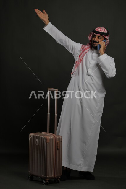  رفع اليد للتعبير عن الوداع والانطلاق لبدء الرحلة، إيماءات الشكر وعدم الحاجه للمساعده، بورتريه لشاب عربي خليجي سعودي مبتسم يرتدي الشماغ والثوب التقليدي يقوم بإجراء مكالمة هاتفية عبر الهاتف المحمول, صورة الجسم بالطول الكامل ، خلفية رمادية