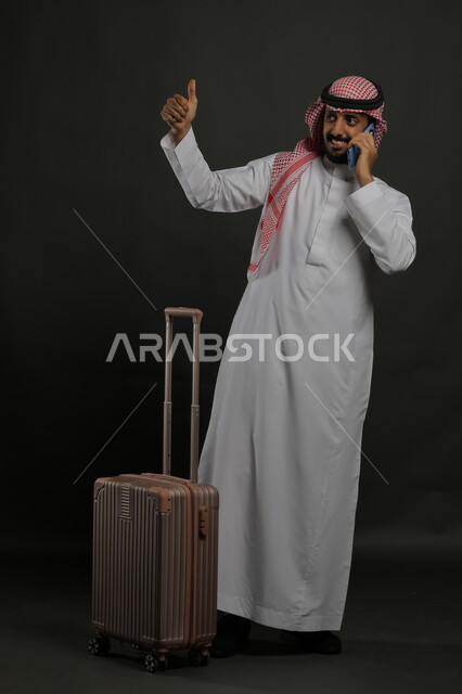 الوداع والانطلاق لبدء الرحلة السياحية بإيماءات القبول والرضا والجودة، الاستمتاع في السياحه بأوقات الفراغ، بورتريه لشاب عربي خليجي سعودي مبتسم يرتدي الشماغ والثوب التقليدي يقومب إجراء مكالمة هاتفية عبر الجوال، صورة الجسم بالطول الكامل ، خلفية رمادية