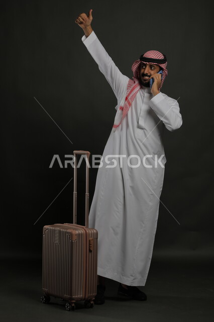 رفع اليد بإيماءات الجوده والتعبير عن الوداع والانطلاق لبدء الرحلة، إيماءات الشكر وعدم الحاجه للمساعده، بورتريه لشاب عربي خليجي سعودي مبتسم يرتدي الشماغ والثوب التقليدي يقوم بإجراء مكالمة هاتفية عبر الهاتف المحمول, صورة الجسم بالطول الكامل ، خلفية رمادية