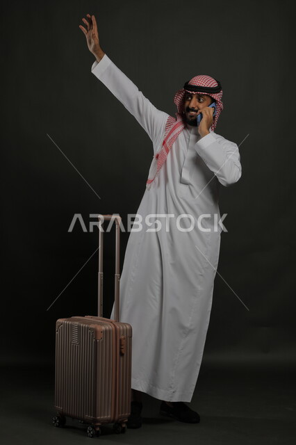 التلويح باليد للتعبير عن الوداع والانطلاق لبدء الرحلة، إيماءات الشكر وعدم الحاجه للمساعده، بورتريه لشاب عربي خليجي سعودي مبتسم يرتدي الشماغ والثوب التقليدي يقوم بإجراء مكالمة هاتفية عبر الهاتف المحمول, صورة الجسم بالطول الكامل ، خلفية رمادية