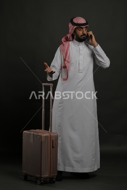 الاستفسار والسؤال عن أمر ما بإيماءات الغضب والقلق, الاستفهام عن خدمات العملاء للرحلة السياحية, بورتريه لرجل عربي خليجي سعودي يرتدي الثوب التقليدي والشماغ يقوم بإجراء مكالمة هاتفية عبر الهاتف المحمول, صورة الجسم بالطول الكامل, خلفية رمادية