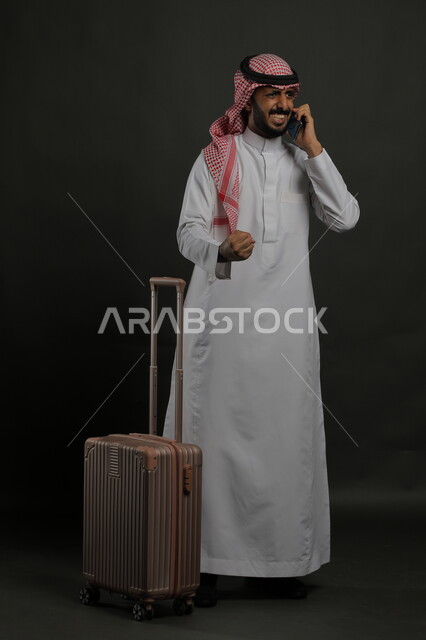 السرور والسعادة وتحقيق الأهداف, بورتريه لرجل عربي خليجي سعودي يرتدي الثوب التقليدي والشماغ يقوم بإجراء مكالمة هاتفية عبر الجوال, البشاشة والفرح لسماع الأخبار الجيده, الاستمتاع بالرحلة السياحية في أوقات العطلة الصيفية, صورة الجسم بالكامل, خلفية رمادية