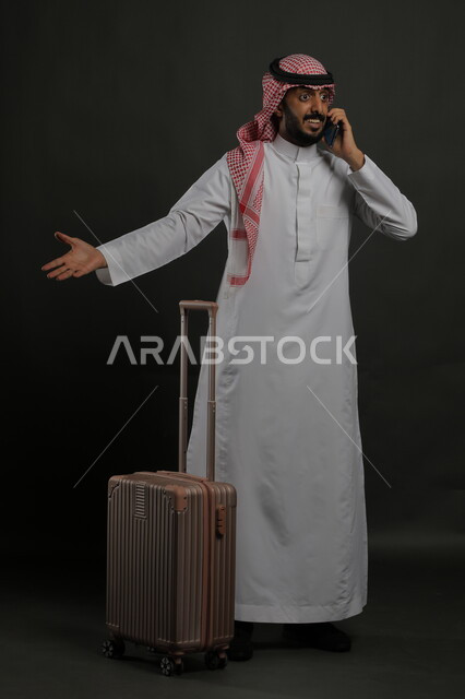 الاستفسار والسؤال عن أمر ما بإيماءات الغضب والاستغراب, الاستفهام عن خدمات العملاء للرحلة السياحية, بورتريه لرجل عربي خليجي سعودي يرتدي الثوب التقليدي والشماغ يقوم بإجراء مكالمة هاتفية عبر الهاتف المحمول, صورة الجسم بالطول الكامل, خلفية رمادية