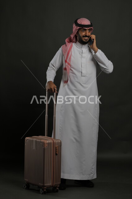 التواصل مع الأهل والأصدقاء, إجراء مكالمات هاتفية عبر الجوال, بورتريه لشاب عربي سعودي خليجي مبتسم يرتدي الثوب التقليدي والشماغ يمسك بيده الهاتف المحمول ويحمل حقيبة السفر, حجز مباشر عبر تطبيقات وبرامج السياحة, صورة الجسم بالطول الكامل, خلفية رمادية