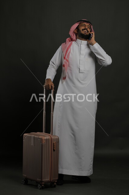 إيماءات السعادة لتحقيق الأهداف, بورتريه لرجل عربي خليجي سعودي مبتسم يرتدي الثوب التقليدي والشماغ يقوم بإجراء مكالمة هاتفية عبر الجوال, البشاشة والفرح لسماع الأخبار الجيده, الاستمتاع بالرحلة السياحية في العطلة الصيفية, صورة الجسم بالكامل, خلفية رمادية