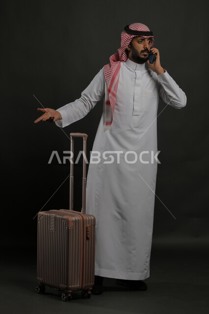 الاستفسار والسؤال عن أمر ما بإيماءات الغضب والقلق, الاستفهام عن خدمات العملاء للرحلة السياحية, بورتريه لرجل عربي خليجي سعودي يرتدي الثوب التقليدي والشماغ يقوم بإجراء مكالمة هاتفية عبر الهاتف المحمول, صورة الجسم بالطول الكامل, خلفية رمادية