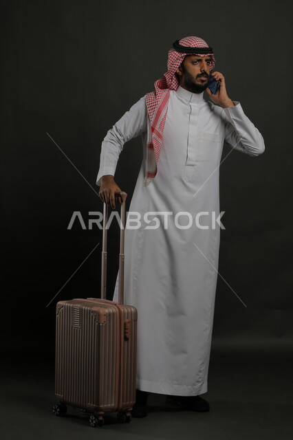 إيماءات الغضب والاستغراب والقلق, التأخر عن موعد الرحلة السياحية, بورتريه لرجل عربي خليجي سعودي يرتدي الثوب التقليدي والشماغ يقوم بإجراء مكالمة هاتفية عبر الهاتف المحمول ويمسك حقيبة السفر, صورة الجسم بالطول الكامل, خلفية رمادية