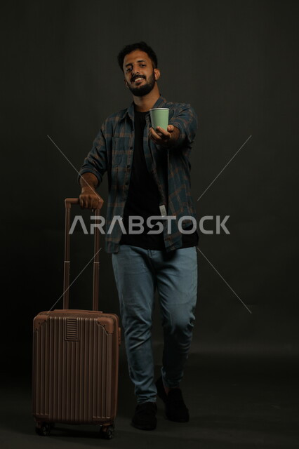 مفهوم الاستعداد  للرحلة السياحية، بورتريه لرجل عربي سعودي خليجي مبتسم يرتدي الزي الكاجوال ويمسك حقيبة السفر، قضاء أوقات مسلية خارج البلاد، احتساء المشروبات الساخنة، صورة الجسم بالطول الكامل، خلفية رمادي