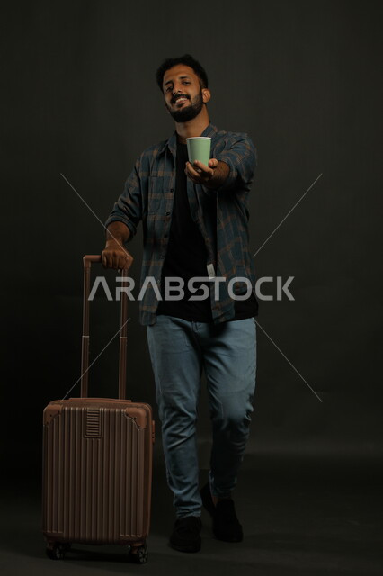 متعة الاستعداد للرحلة السياحية، بورتريه لشاب عربي سعودي خليجي مبتسم يرتدي الزي الكاجوال ويمسك حقيبة السفر، قضاء أوقات مسلية خارج البلاد، احتساء المشروبات الساخنة، صورة الجسم بالكامل، خلفية رمادي