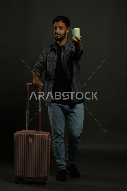 الاستعداد لمغادرة البلاد, قضاء عطلة صيفية ممتعة, بورتريه لرجل عربي سعودي خليجي مبتسم يرتدي الزي الكاجوال يمسك بيده حقيبة السفر, تقديم المشروبات الساخة, صورة لكامل الجسم, خلفية رمادية
