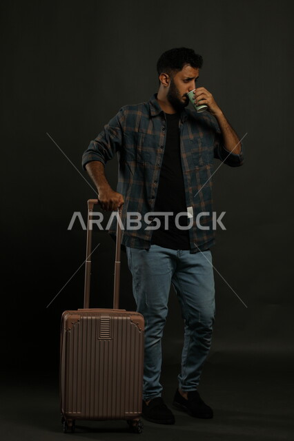 Spending fun times outside the country, portrait of a smiling Saudi Gulf Arab man wearing casual clothes and holding a suitcase and a hot cup of coffee, concept of tourist trips, gestures of enthusiasm for an entertaining trip, full-length body portrait, gray background