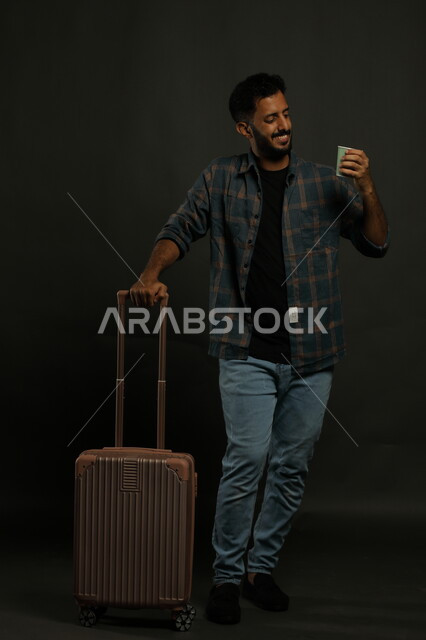 الإستمتاع بالمشروبات الدافئة، بورتريه  لرجل عربي سعودي خليجي مبتسم يرتدي الزي الكاجوال ويحمل حقيبة السفر، الابتعاد عن ضغوط الحياة، استكشاف ثقافات جديدة، الإستعداد للرحلة السياحية، صورة الجسم بالطول الكامل، خلفية رمادي