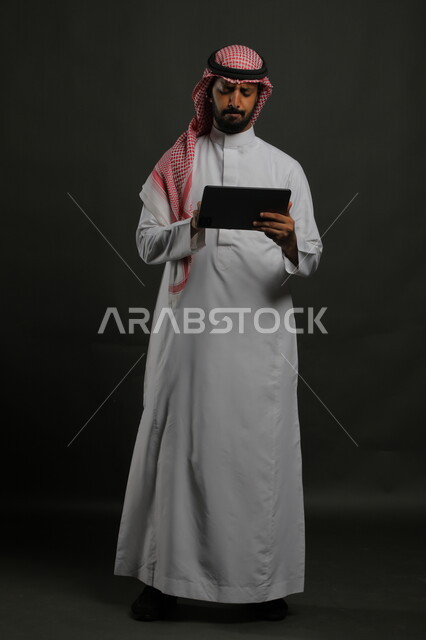 قراءة خبر غير سار على مواقع الانترنت، استخدام التقنيات الحديثة والمطورة، بورتريه لرجل عربي خليجي سعودي يرتدي الزي التقليدي يحمل بيده جهاز التابلت، إيماءات الانزعاج والغضب أثناء اللعب على تطبيقات الأجهزة اللوحية، صورة الجسم بالكامل, خلفية سوداء