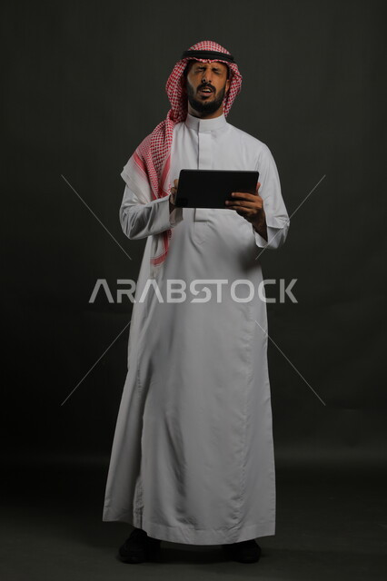 قراءة خبر غير سار على مواقع الانترنت، استخدام التقنيات الحديثة والمطورة، بورتريه لرجل عربي خليجي سعودي يرتدي الزي التقليدي يحمل بيده جهاز التابلت، إيماءات الانزعاج والغضب أثناء اللعب على تطبيقات الأجهزة اللوحية، صورة الجسم بالكامل, خلفية سوداء