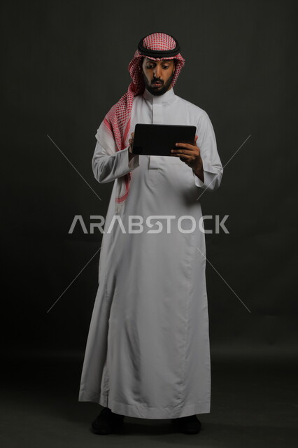 ايماءات الانفعال والاندماج أثناء مشاهدة  شيء ما على الجهاز اللوحي، الحماس والدهشه بالفوز والنصر، بورتريه لرجل عربي خليجي سعودي يرتدي الزي التقليدي يقوم بإجراء مكالمة فيديو وسماع خبر مفاجئ ،صورة الجسم بالطول الكامل، خلفية سوداء