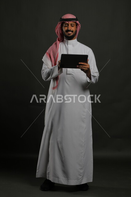 استخدام التقنيات الحديثة المتطورة, بورتريه لرجل عربي خليجي سعودي مبتسم يرتدي الزي التقليدي يحمل بيده جهاز التابليت، الدردشة مع الأهل والأصدقاء, متابعة آخر أخبار السوشال ميديا، تطبيقات وبرامج الأجهزة اللوحية، صورة الجسم بالكامل, خلفية سوداء