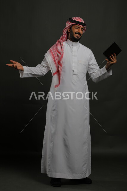 Watching comedic and funny movies on the Internet, making a video call with family and friends, a portrait of a smiling Saudi Gulf Arab man wearing traditional clothing holding a tablet in his hand with gestures of happiness and pleasure, full body portrait, black background