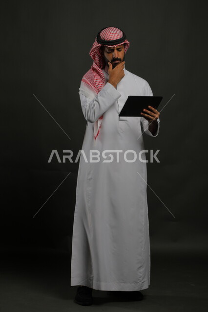 Gestures of annoyance and anger while using tablet applications, reading unpleasant news on websites, using modern and developed technologies, portrait of a Saudi Gulf Arab man wearing traditional clothing holding a tablet in his hand, full-length body image, black background